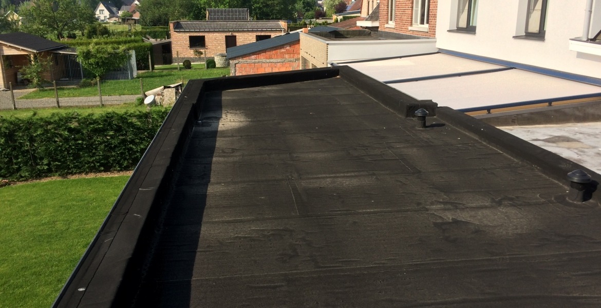 Czarny, płaski dach pokryty papą termozgrzewalną z widocznymi kominkami wentylacyjnymi, w tle zabudowa jednorodzinna i zieleń.