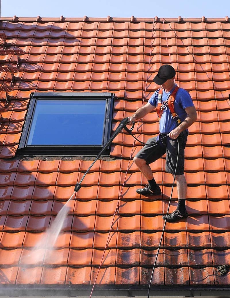 Pracownik w uprzęży, z czapką na głowie, czyści czerwoną dachówkę z okienkiem dachowym za pomocą myjki ciśnieniowej, strumień wody usuwa zabrudzenia.