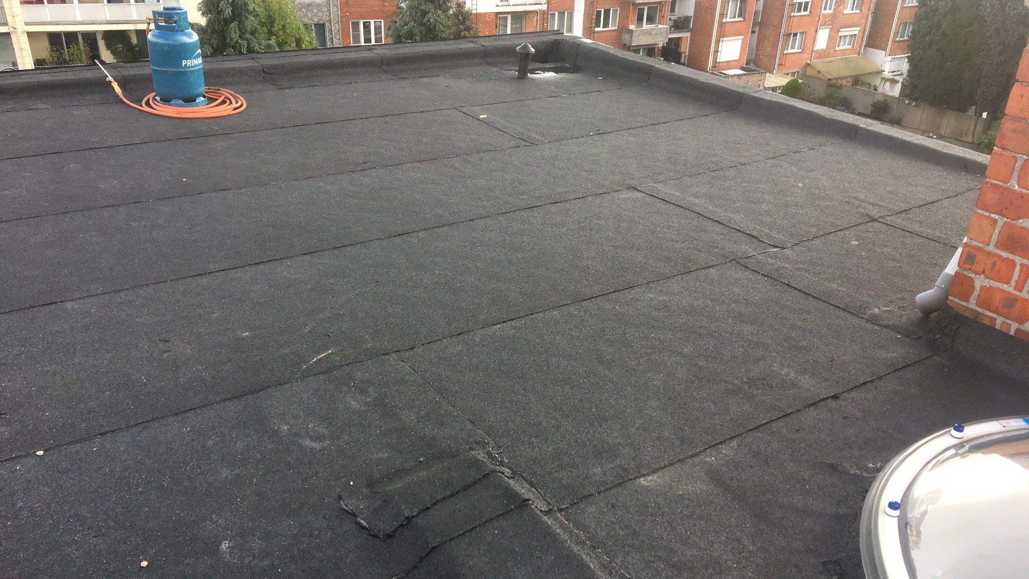 Wykończony, płaski dach pokryty czarną papą termozgrzewalną w Warszawie, widoczny komin wentylacyjny, butla gazowa z palnikiem oraz fragment ściany budynku z rynną spustową.