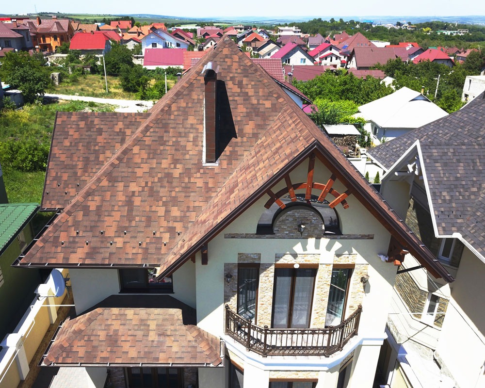 Widok z góry na dom z brązowym dachem z gontu bitumicznego, widoczny komin, balkon z kutą balustradą i dekoracyjne elementy drewniane na szczycie, w tle inne domy.