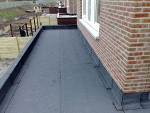 Wykończony płaski dach pokryty czarną papą termozgrzewalną, przylegający do ściany z czerwonej cegły. Widoczny fragment okna i drewniane elementy konstrukcyjne. Ujęcie z perspektywy dachu, w tle...