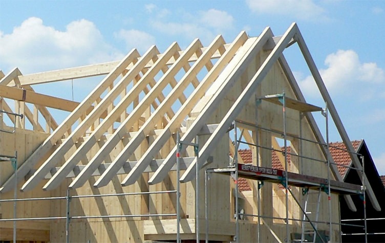 Konstrukcja drewniana dachu w trakcie budowy, widoczne krokwie i ściany szkieletowe, rusztowanie przy ścianie budynku, błękitne niebo w tle.