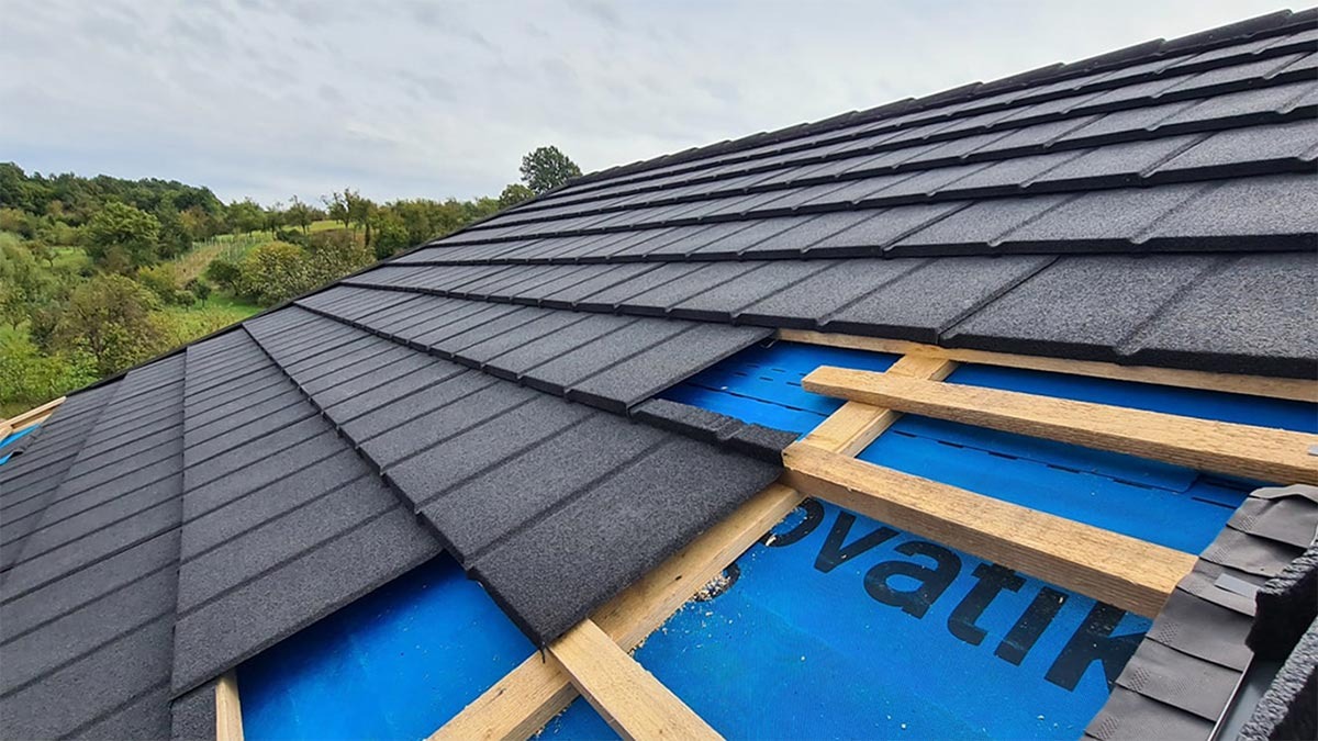 Montaż grafitowej blachodachówki modułowej na dachu z widoczną membraną i elementami drewnianej konstrukcji. W tle zielony krajobraz.