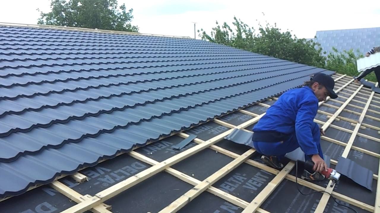 Monter w niebieskim kombinezonie przycina blachodachówkę szlifierką kątową na drewnianej konstrukcji dachu pokrytej czarną membraną, w tle widoczny fragment gotowego pokrycia.