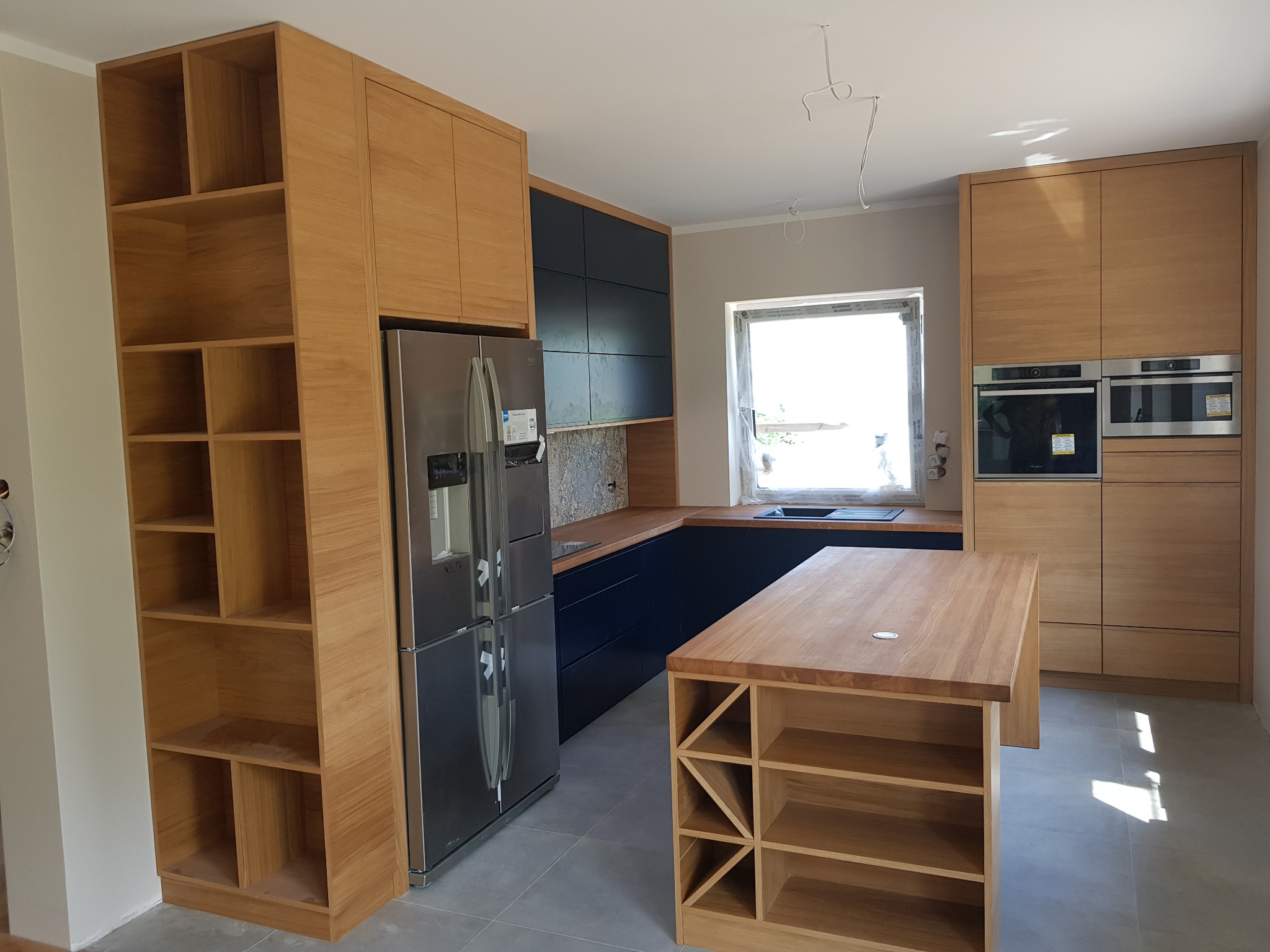 Nowoczesna kuchnia z drewnianymi frontami w odcieniach dębu, ciemnogranatowymi szafkami dolnymi i wyspą z półkami na wino, uzupełniona o srebrną lodówkę i dwa piekarniki w zabudowie.