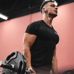 Mężczyzna w czarnej koszulce z słuchawkami, podnosi sztangę w siłowni, widoczny z dołu, oświetlenie górne.