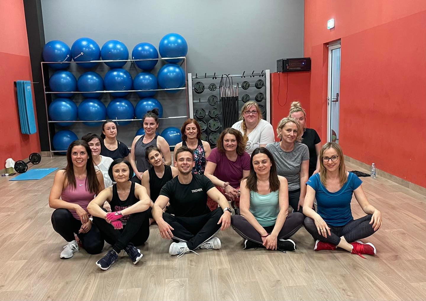 Grupa kobiet i mężczyzna z napisem 'TRENER' na koszulce siedząca po turecku na podłodze sali gimnastycznej, za nimi rząd niebieskich piłek do ćwiczeń i sprzęt do ćwiczeń.