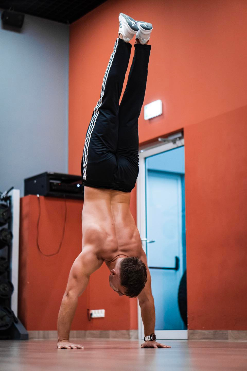 Mężczyzna w czarnych spodniach z białymi paskami i białych butach robiący stójkę w pomieszczeniu z pomarańczową ścianą.