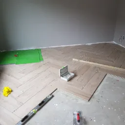 Układanie podłogi z płytek imitujących drewno w jodełkę, widoczny poziomica, skrzynka z krzyżykami dystansowymi, żółta ściereczka i fragmenty niezakończonej podłogi.