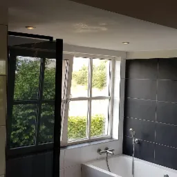 Nowoczesna łazienka z czarnymi płytkami na ścianie, białą wanną i oknem z widokiem na zieleń, z widocznym czarnym panelem prysznicowym.