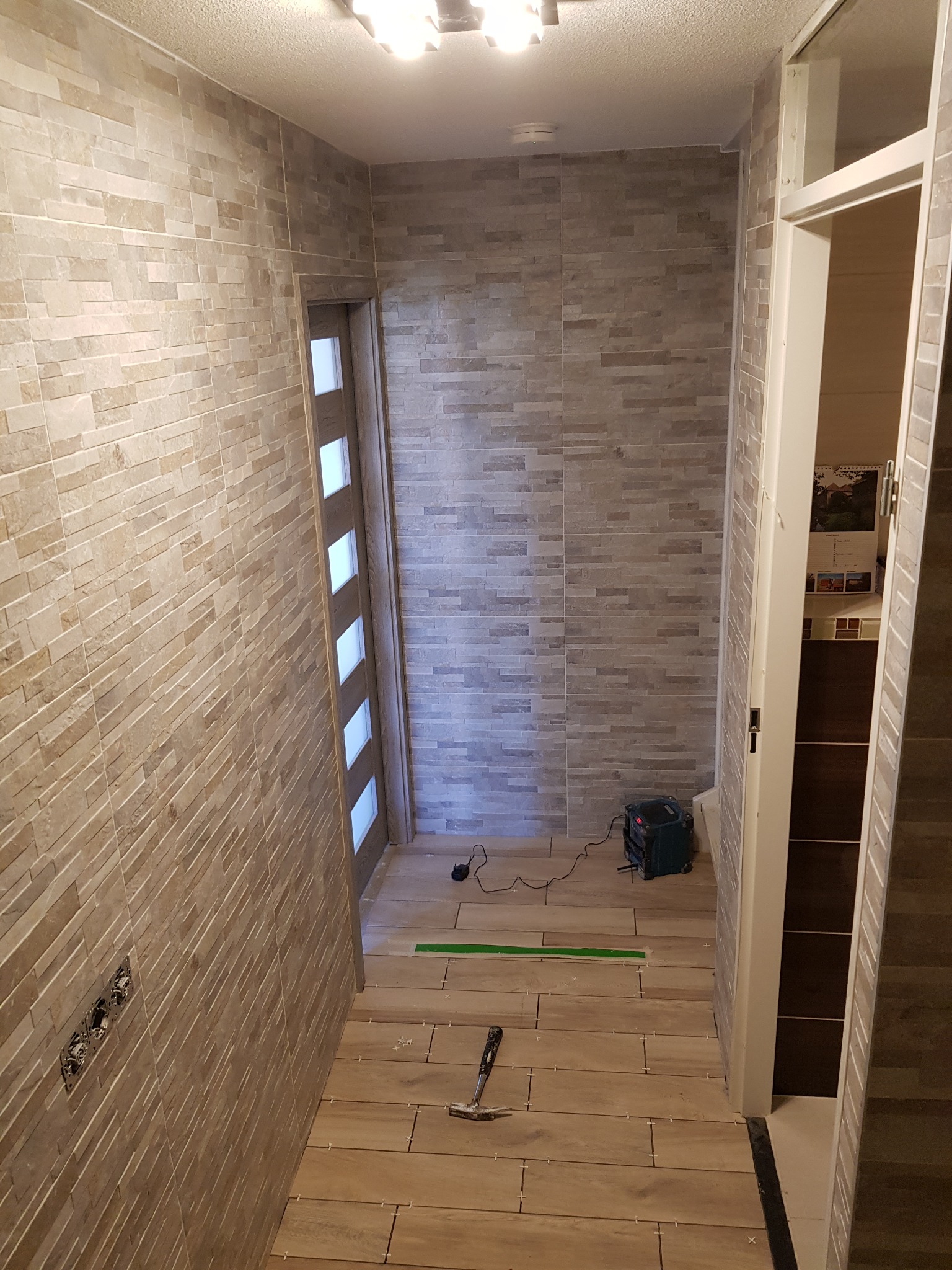 Wąski korytarz w trakcie wykańczania: ułożone płytki podłogowe w odcieniach beżu, ściany wyłożone szarobrązową mozaiką, widoczne krzyżyki dystansowe, młotek i osuszacz powietrza.