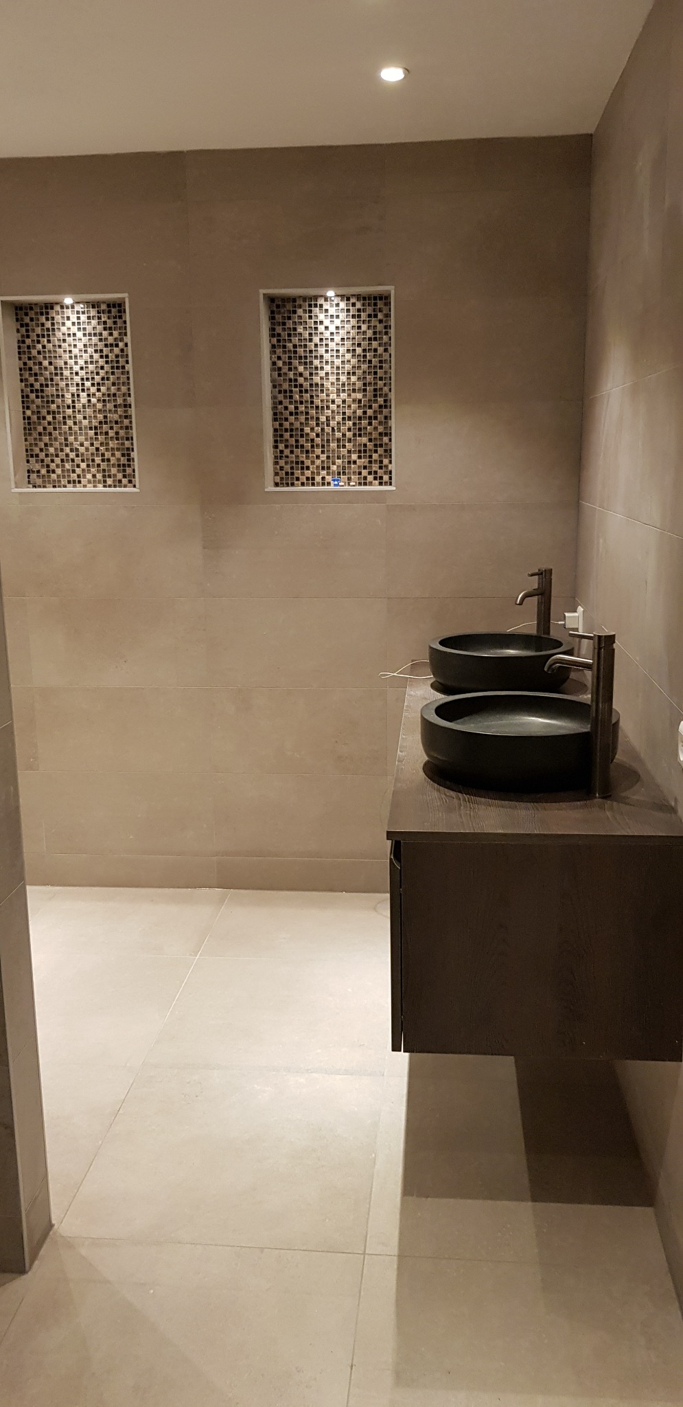Nowoczesna łazienka z dwoma czarnymi umywalkami w kształcie misy na drewnianej szafce, beżowe płytki na ścianach i podłodze oraz dekoracyjne wnęki z mozaiką.