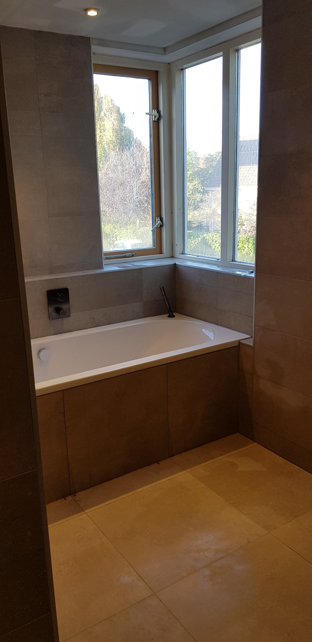 Wykończona łazienka z wanną obudowaną panelami w kolorze płytek, okno z widokiem na drzewa, szare płytki na ścianach i podłodze.