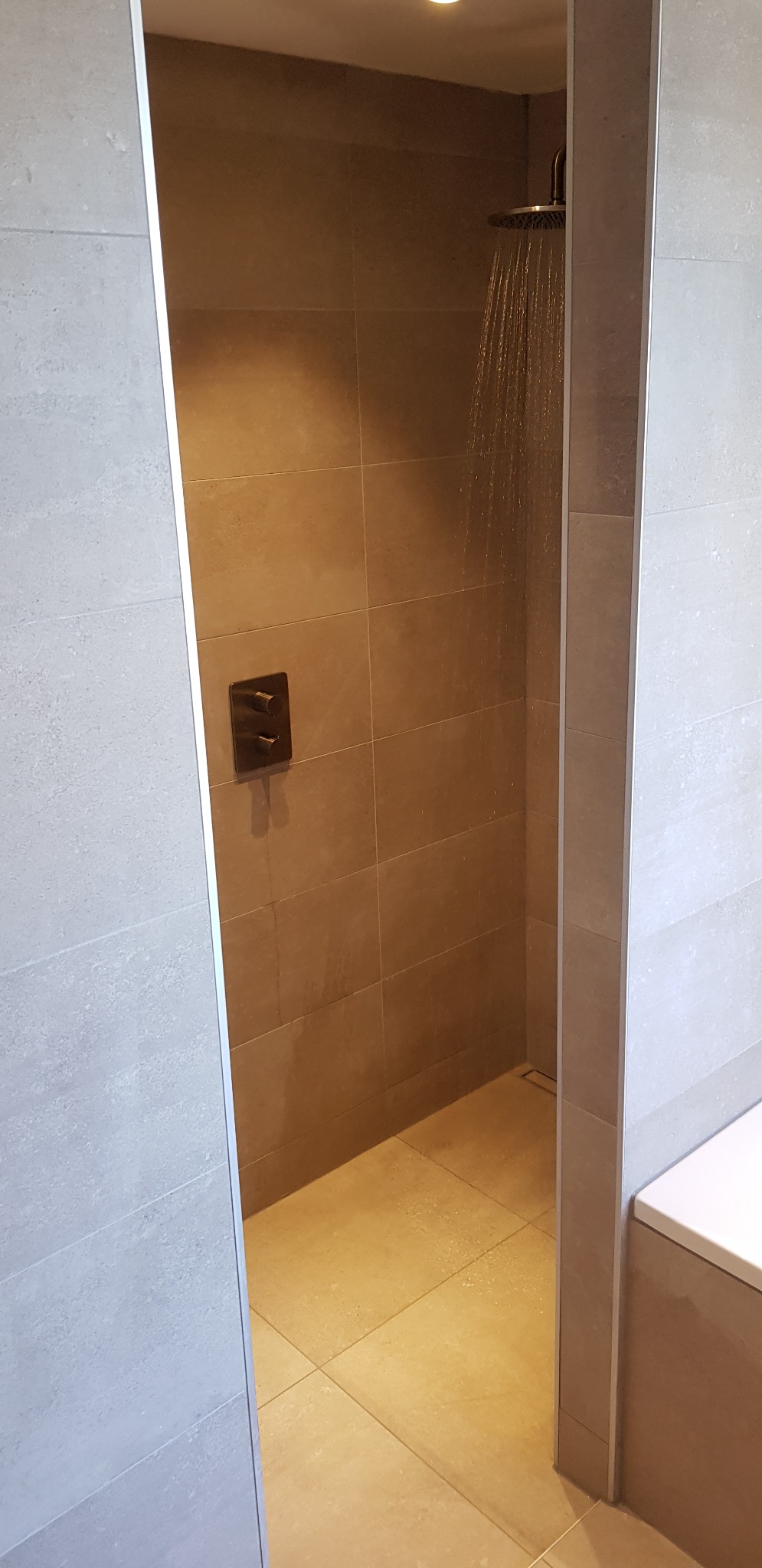 Nowoczesny prysznic typu walk-in z deszczownicą w trakcie użycia, widoczne płytki w odcieniach beżu i szarości oraz termostat natynkowy w ciemnym kolorze.