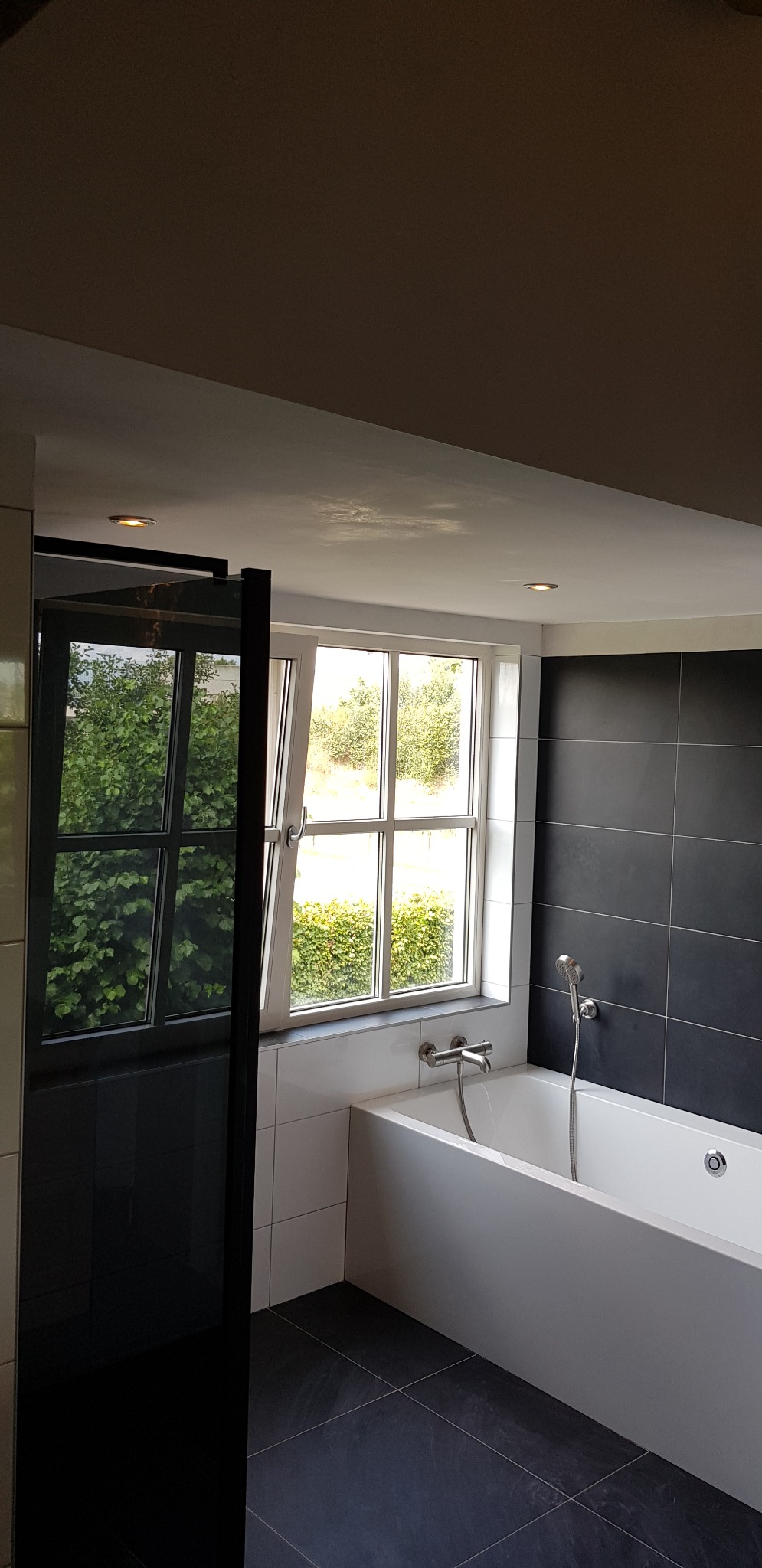 Nowoczesna łazienka z czarnymi płytkami na ścianie, białą wanną i oknem z widokiem na zieleń, z widocznym czarnym panelem prysznicowym.