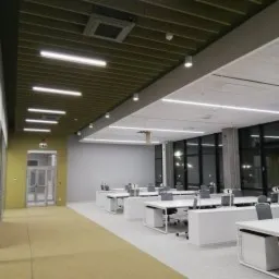 Nowoczesne biuro z białymi biurkami i oświetleniem LED w suficie podwieszanym, widok z korytarza z wykładziną dywanową.