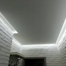 Białe, matowe wykończenie sufitu z ukrytym oświetleniem LED, ściany wyłożone białą cegłą dekoracyjną, widoczny fragment wieszaka z haczykami.