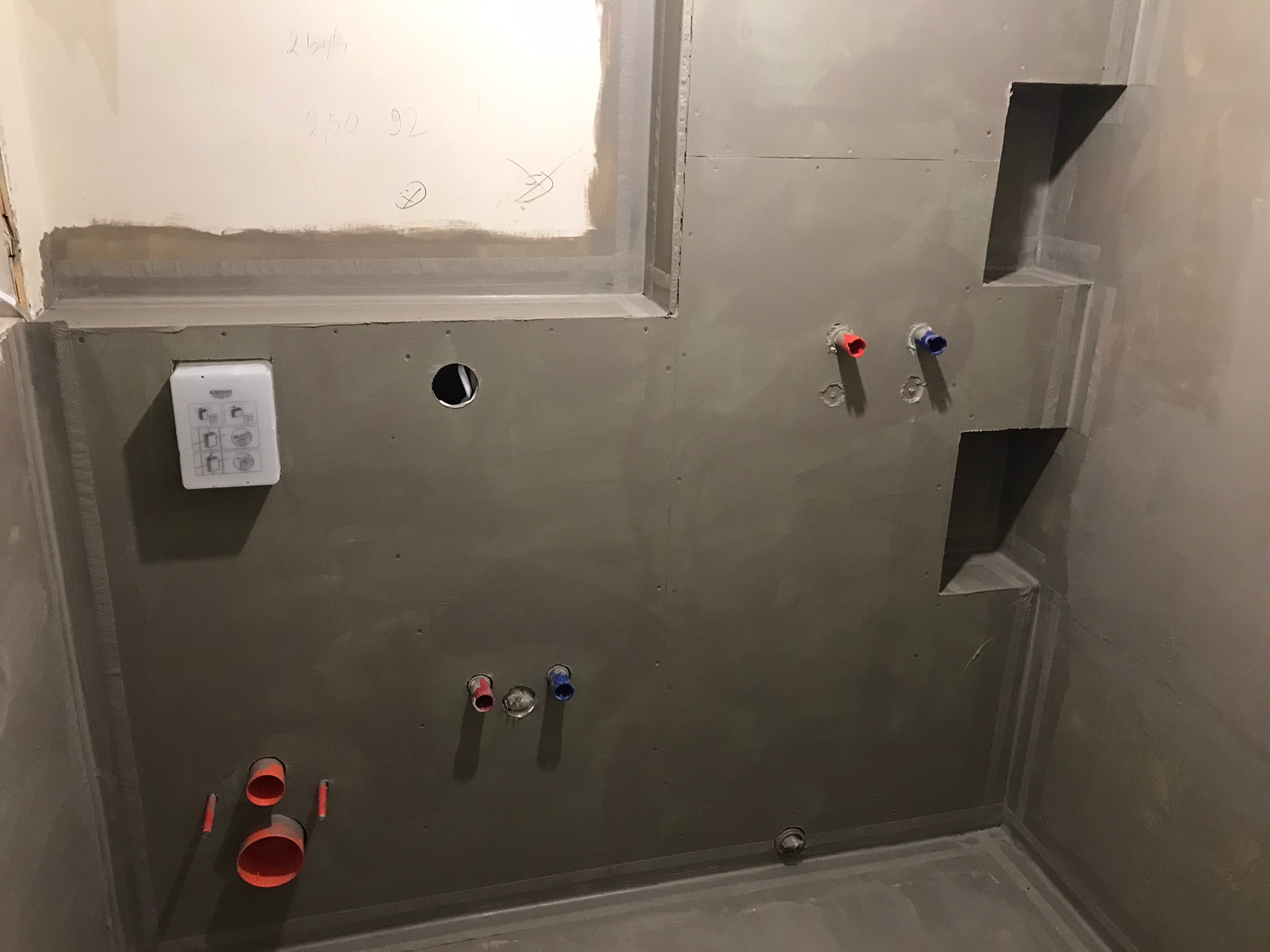 Przygotowana do kafelkowania ściana w łazience z wnękami, widoczne wyprowadzenia instalacji wodnej, zabudowa Grohe i ślady ołówka na ścianie.