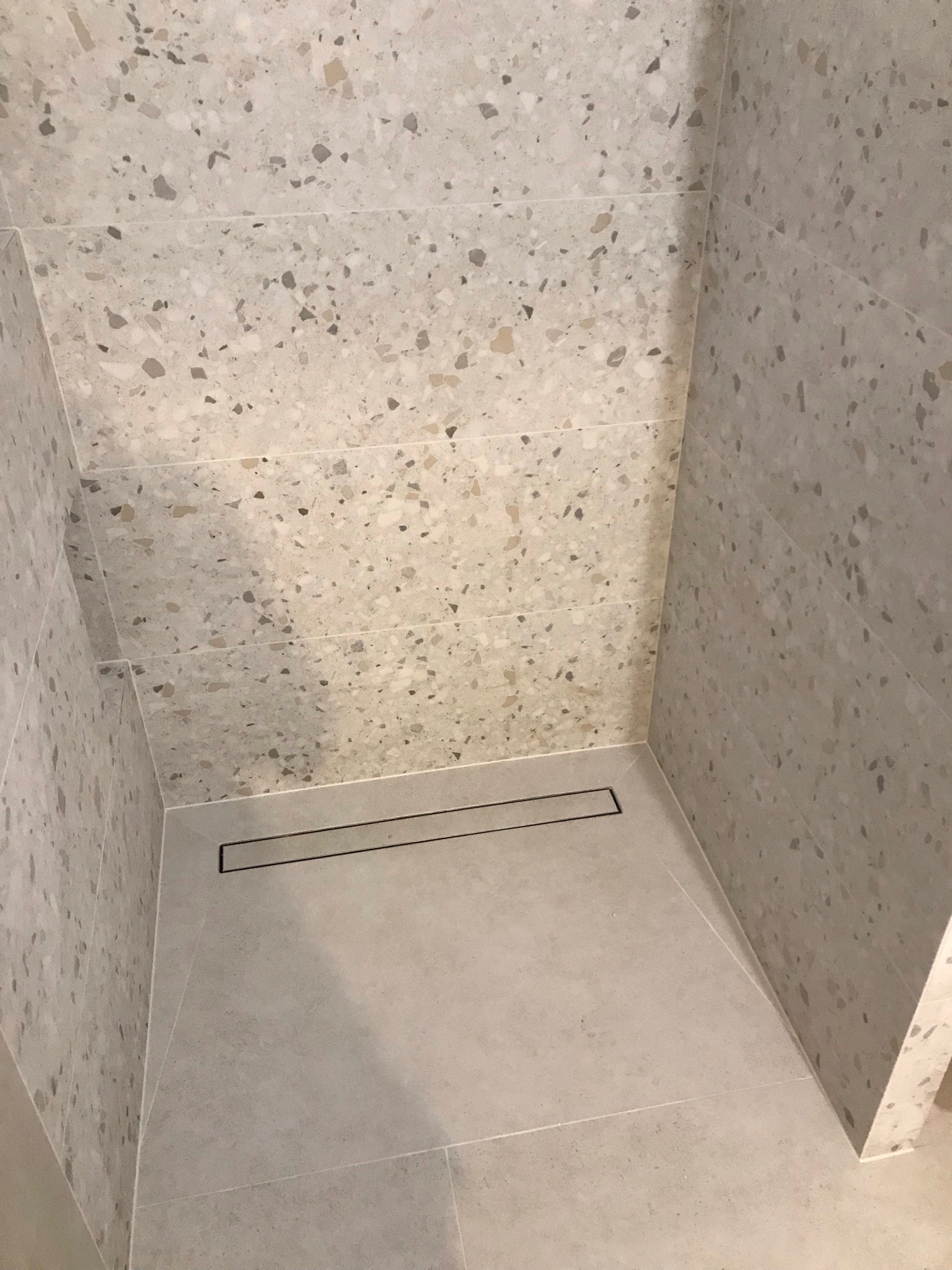 Nowoczesny prysznic walk-in z odpływem liniowym, wykończony płytkami imitującymi lastryko w odcieniach szarości i beżu.