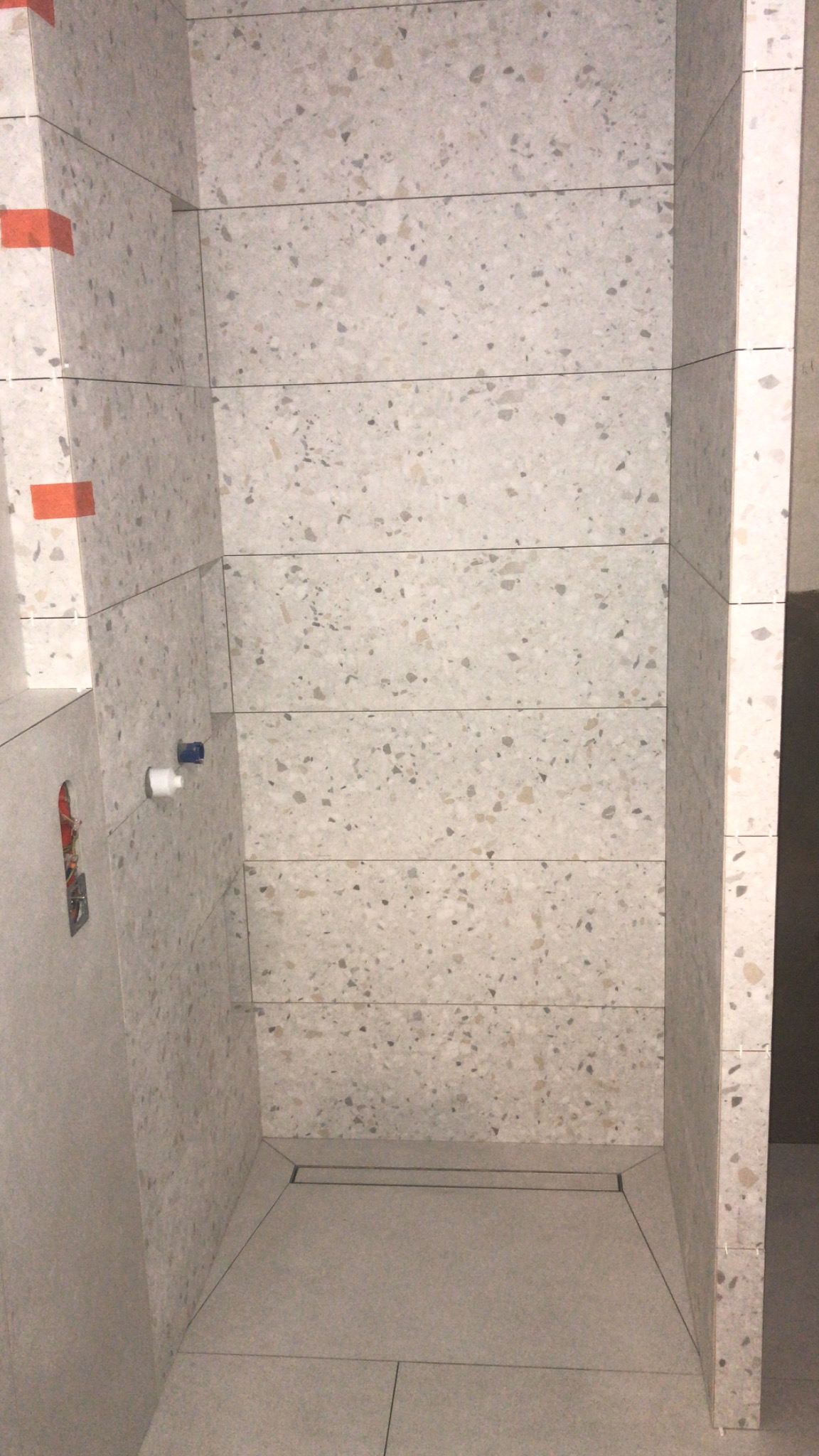 Wykończony brodzik kabiny prysznicowej z odpływem liniowym, wyłożony płytkami imitującymi lastryko, z widocznymi przyłączami wodnymi i pomarańczowymi taśmami maskującymi na fugach.