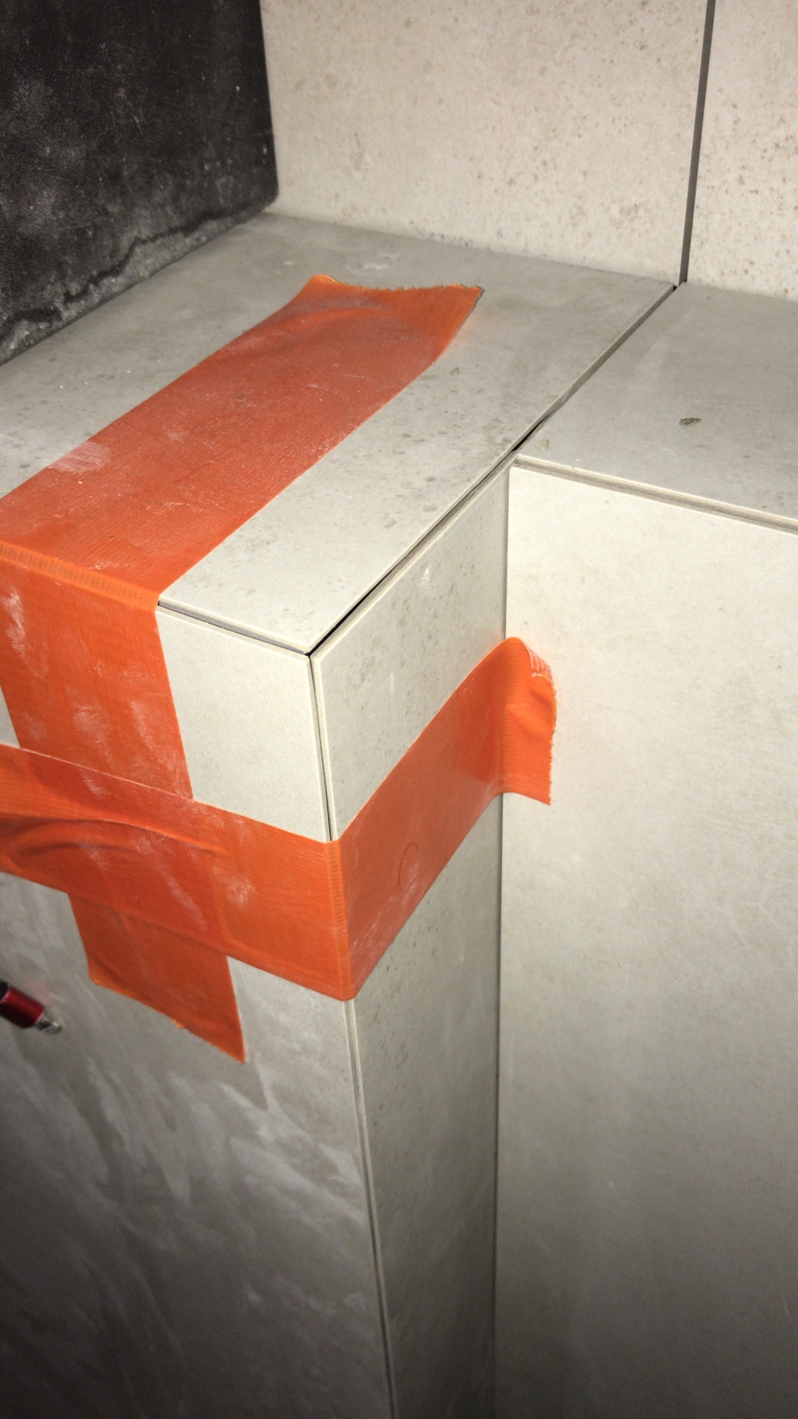 Szczegółowe ujęcie narożnika wykończonego jasnymi płytkami, zabezpieczonego pomarańczową taśmą malarską, widoczny fragment ściany w tle.