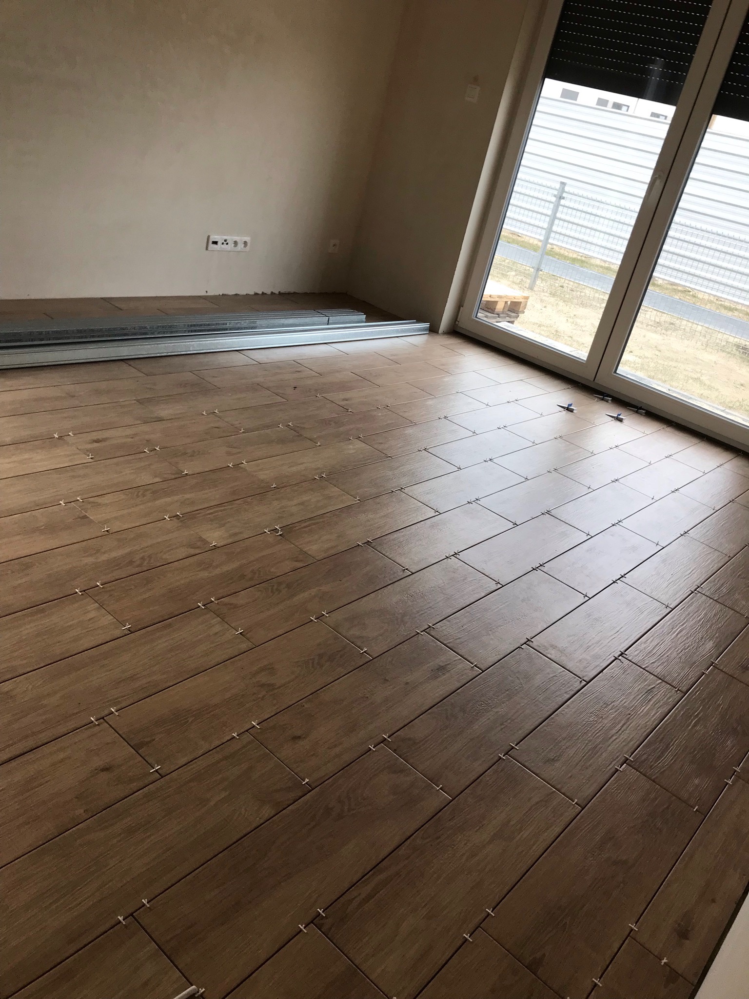 Ułożona podłoga z płytek imitujących drewno w nowym pomieszczeniu, widoczne krzyżyki dystansowe, profile metalowe przy ścianie i okno z roletami zewnętrznymi.