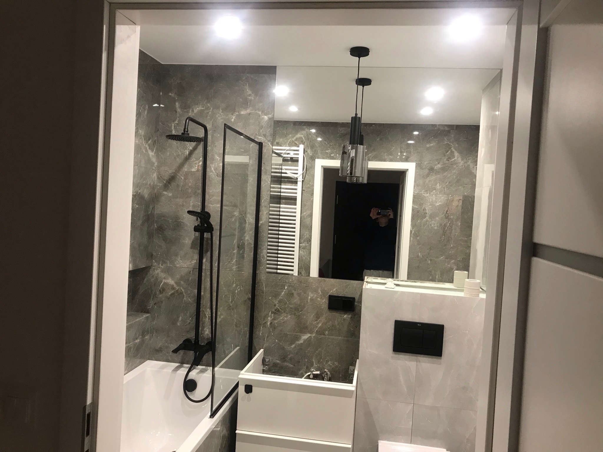 Wykończona łazienka z szarymi marmurkowymi płytkami, czarną armaturą, minimalistyczną lampą wiszącą i widocznym odbiciem fotografa w lustrze.