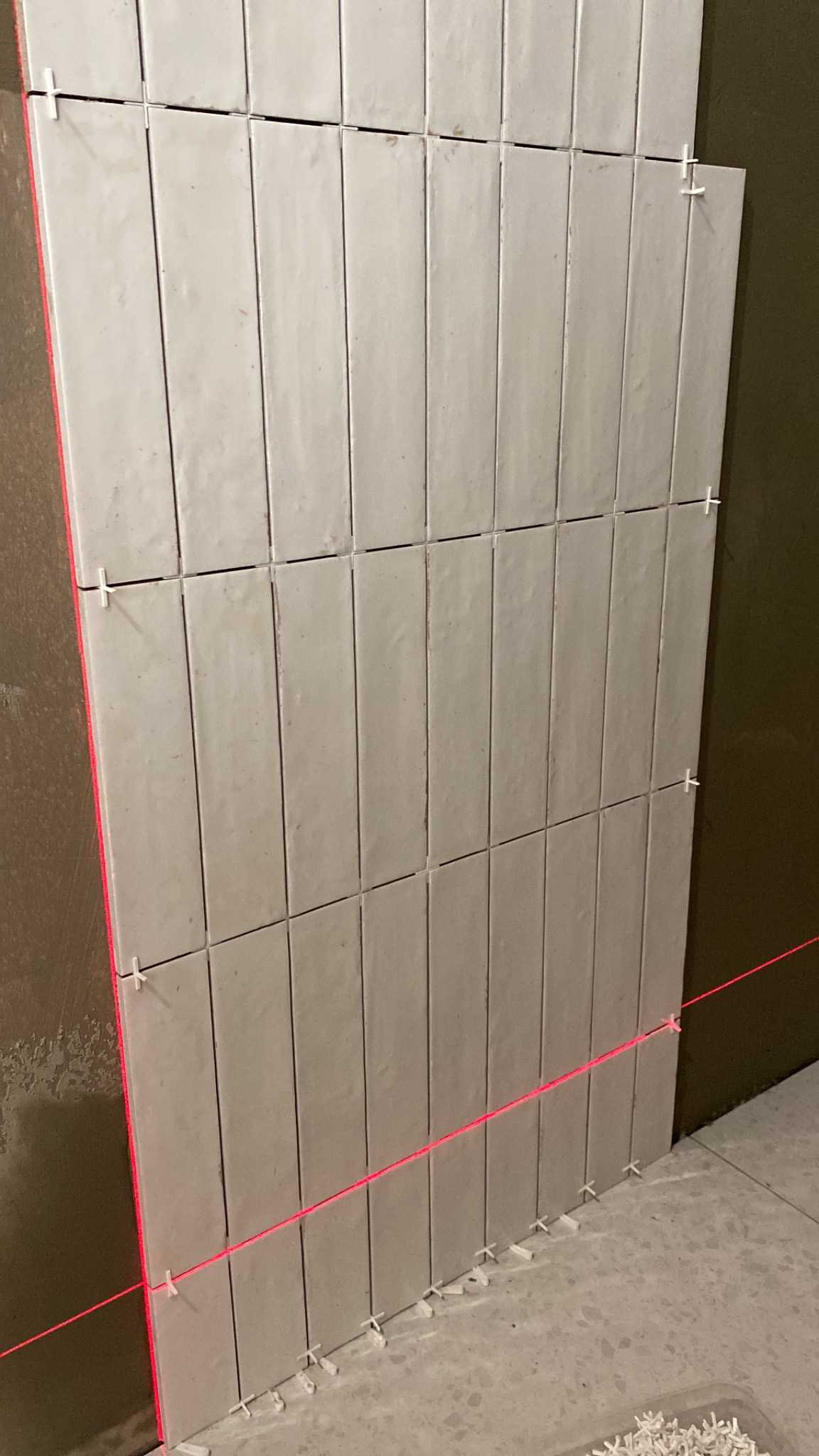 Układanie podłużnych, matowych płytek na ścianie z użyciem poziomicy laserowej i krzyżyków dystansowych.