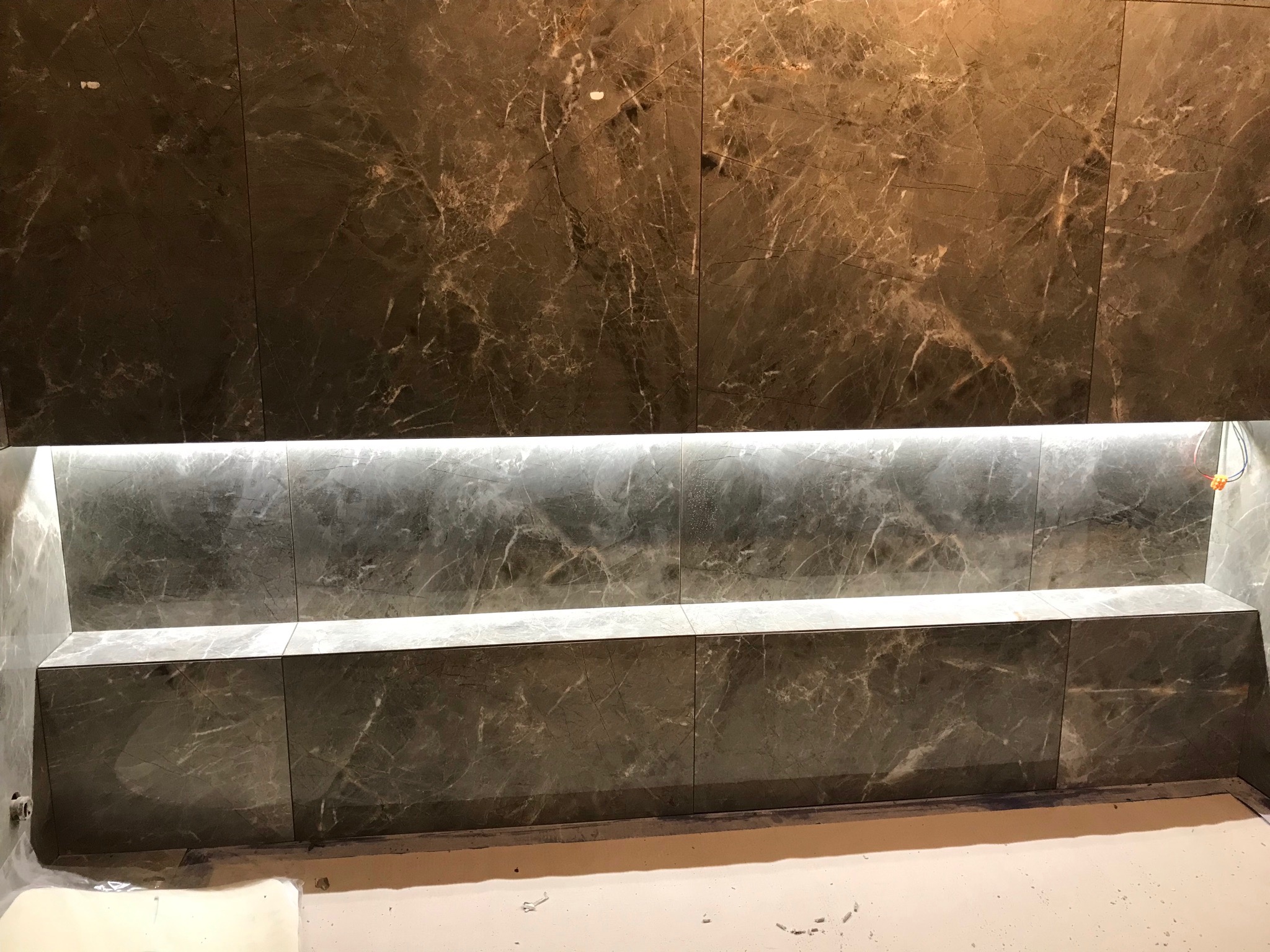 Ściana i półka wyłożone brązowymi płytkami imitującymi marmur z podświetleniem LED, widoczne niedociągnięcia na podłodze.