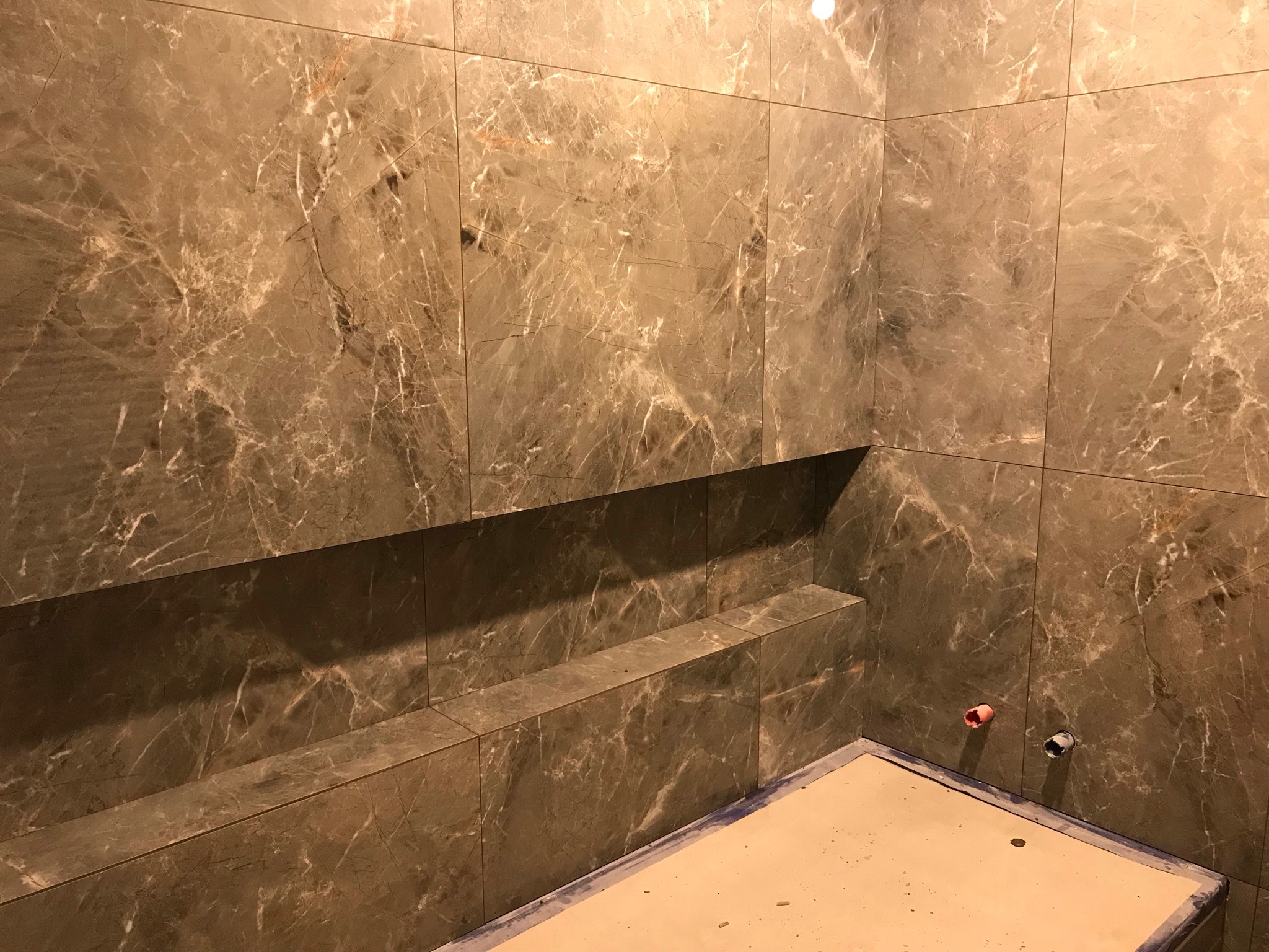 Wnętrze łazienki w trakcie wykańczania, ściany i półki wyłożone płytkami imitującymi marmur w odcieniach szarości i brązu, widoczne rury instalacyjne wystające ze ściany, podłoga zabezpieczona...