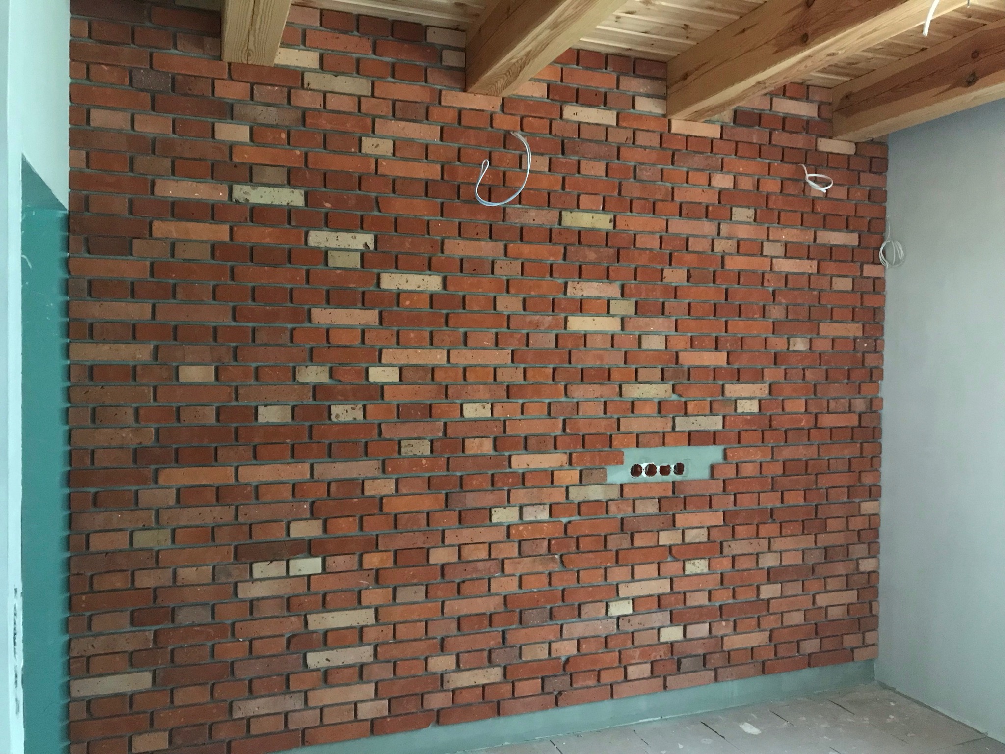 Ściana z cegły klinkierowej w odcieniach czerwieni i beżu, z widocznymi przewodami elektrycznymi i przygotowanym miejscem na gniazdka, sufit z drewnianych belek.