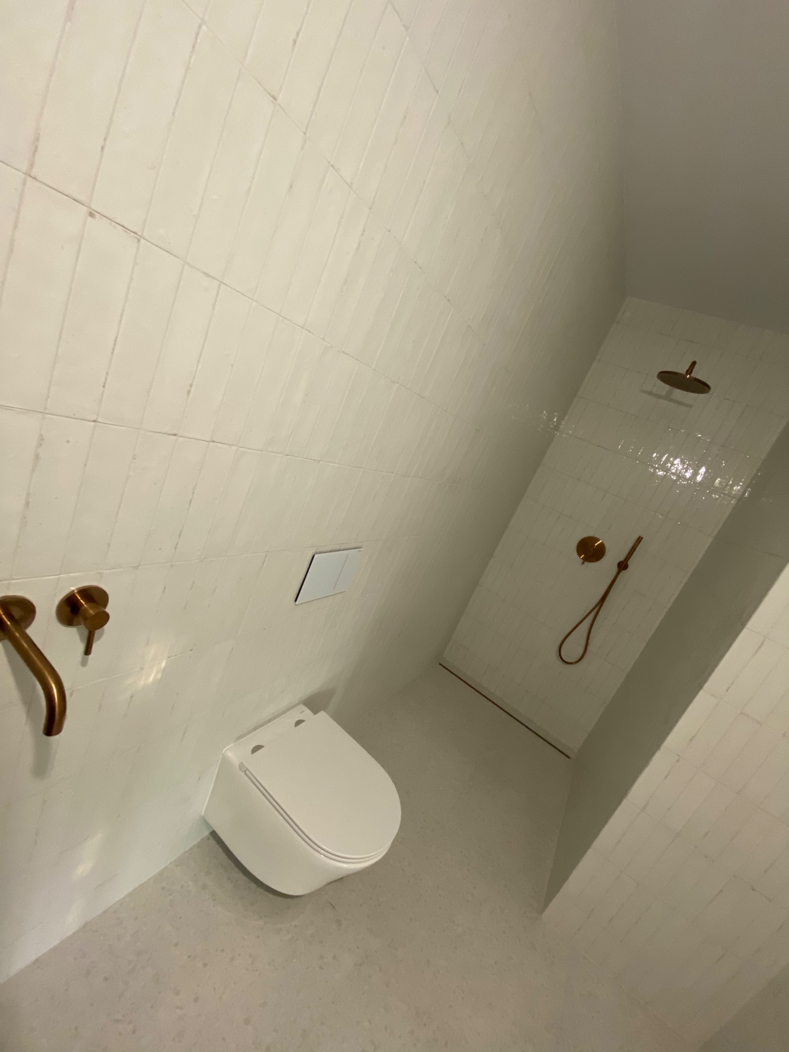 Biała łazienka z toaletą podwieszaną, armaturą w kolorze mosiądzu i kabiną prysznicową typu walk-in wyłożona wąskimi płytkami.