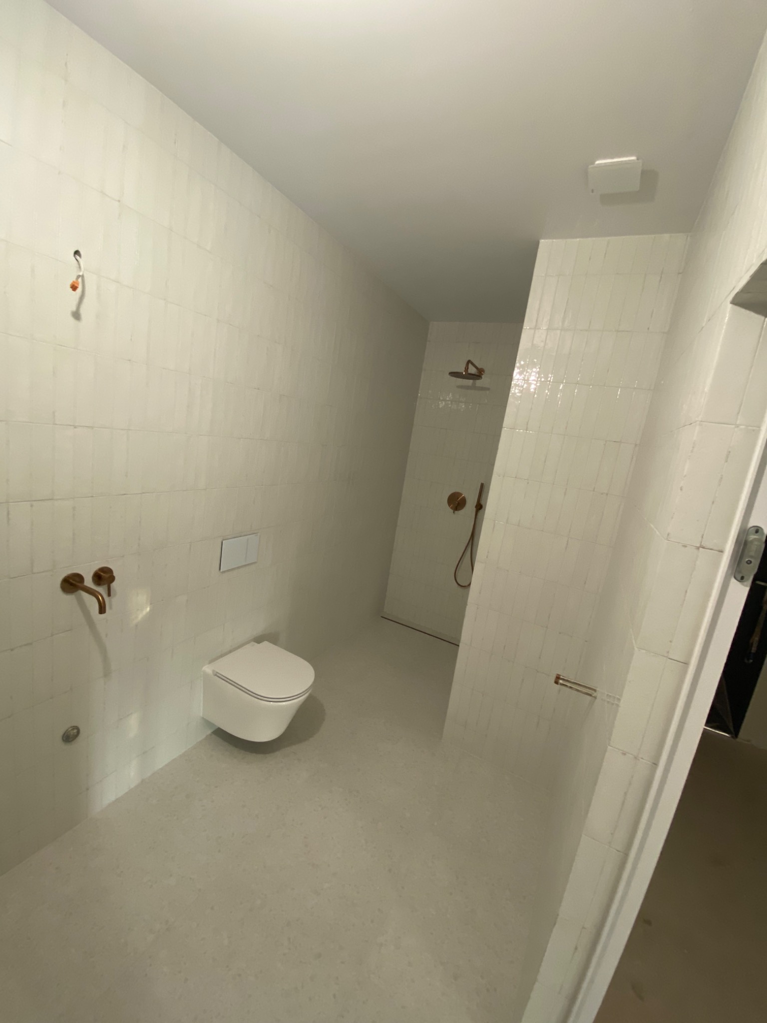 Nowoczesna łazienka z białymi, wąskimi płytkami ułożonymi pionowo, armatura w kolorze miedzi, podwieszana toaleta, widoczne niedokończone podłączenie elektryczne po lewej stronie.