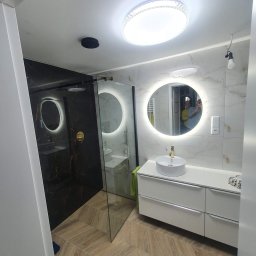 Karbud - Nowoczesna łazienka z kabiną prysznicową typu walk-in, umywalką nablatową na białej szafce, okrągłym lustrem z podświetleniem LED oraz podłogą ułożoną w jodełkę.