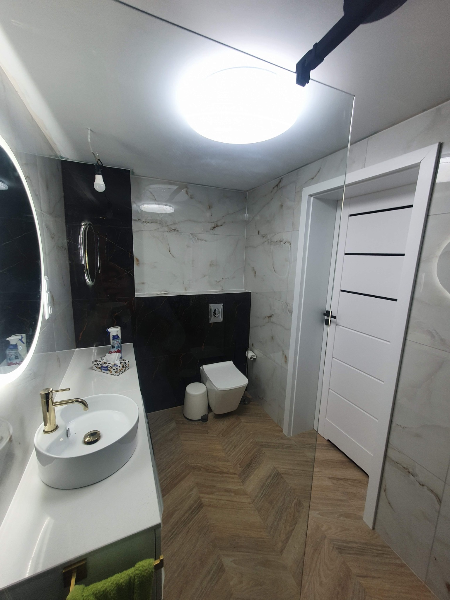 Nowoczesna łazienka z umywalką nablatową, złotą armaturą, lustrem z podświetleniem LED, płytkami imitującymi marmur, oraz drewnopodobną podłogą ułożoną w jodełkę.