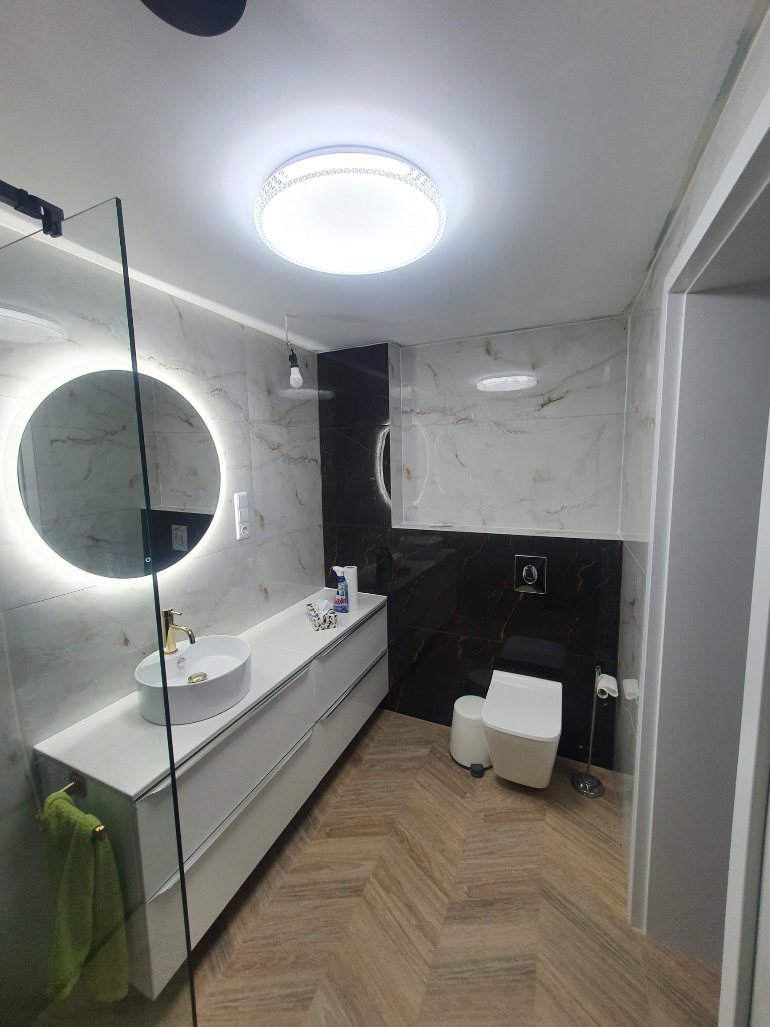 Nowoczesna łazienka z marmurowymi płytkami, okrągłym podświetlanym lustrem i drewnopodobną podłogą ułożoną w jodełkę, widok na umywalkę z złotą armaturą, toaletę i szafkę podumywalkową.