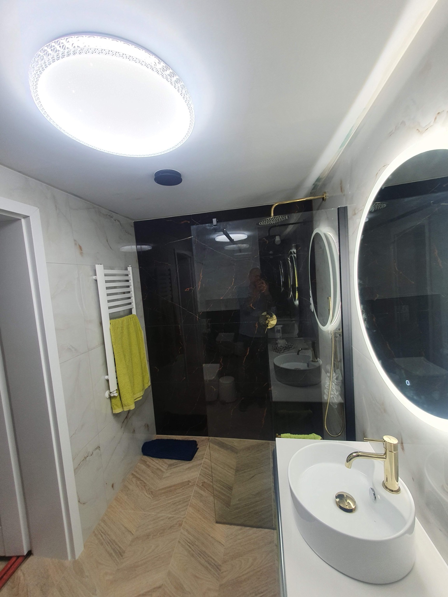 Nowoczesna łazienka z owalnym lustrem z podświetleniem LED, kabiną prysznicową typu walk-in z czarnymi marmurowymi panelami, białą umywalką nablatową z złotą armaturą i grzejnikiem drabinkowym...