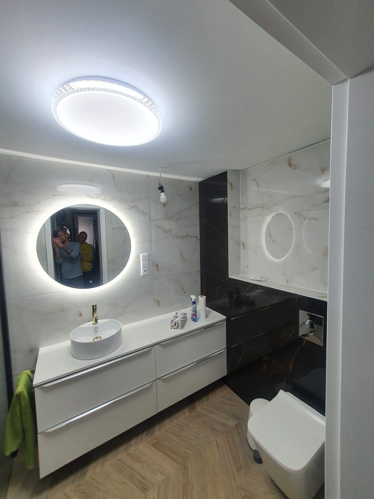 Nowoczesna łazienka z białymi marmurkowymi płytkami, podświetlanym lustrem, białą umywalką nablatową i szafką z szufladami, oraz toaletą w zabudowie; odbicie dwóch osób w lustrze.