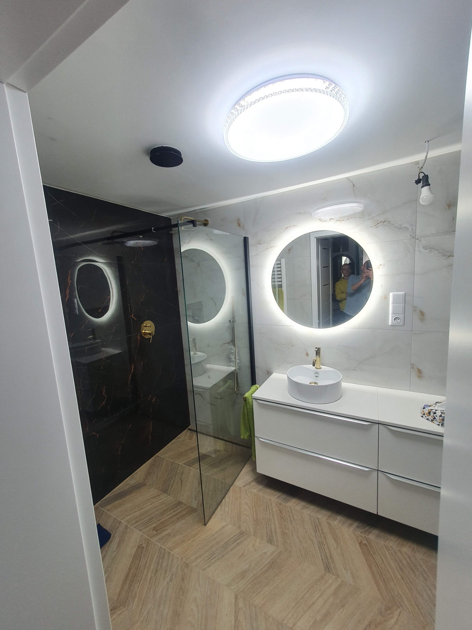 Nowoczesna łazienka z kabiną prysznicową typu walk-in, umywalką nablatową na białej szafce, okrągłym lustrem z podświetleniem LED oraz podłogą ułożoną w jodełkę.