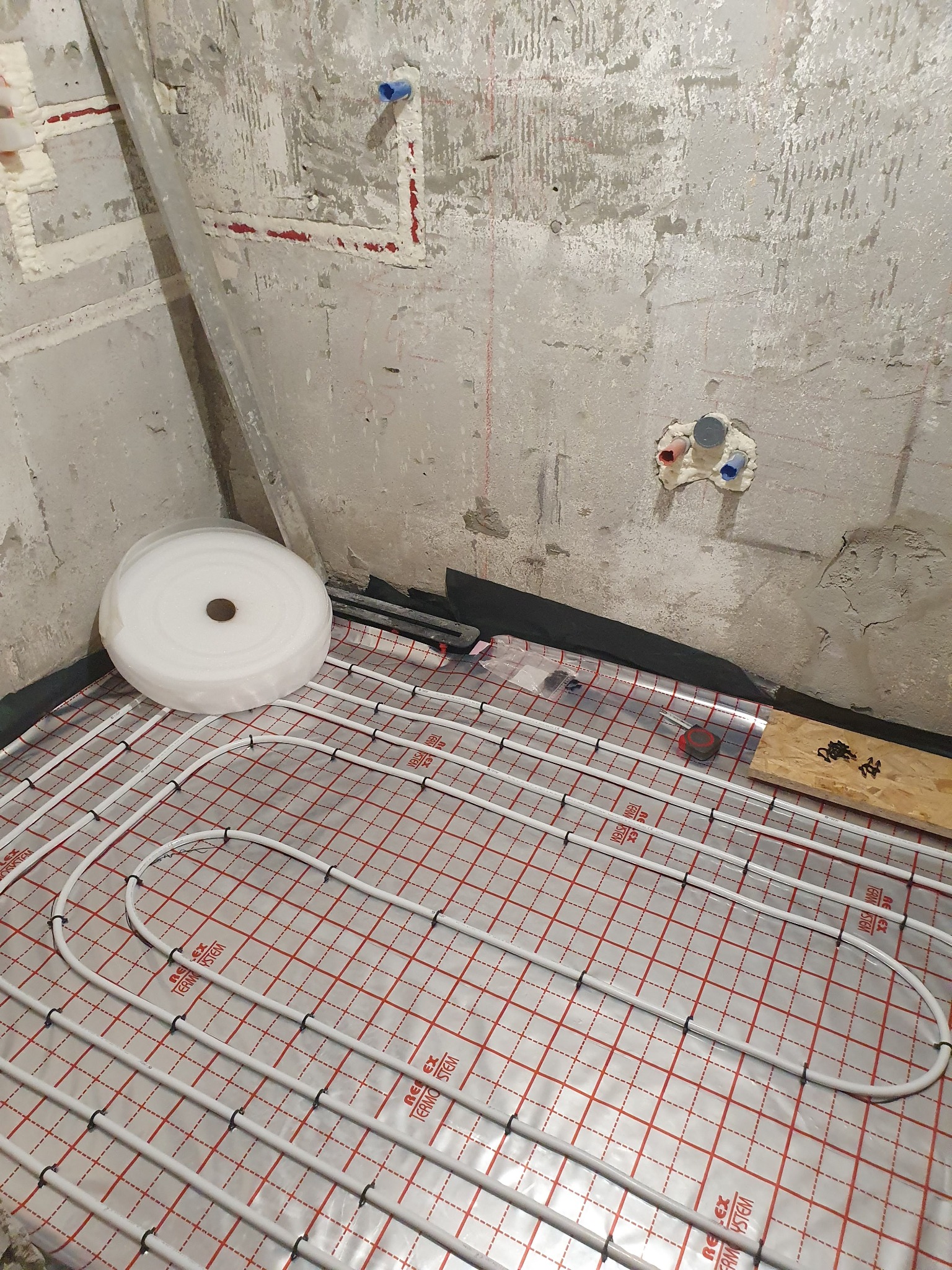 Instalacja ogrzewania podłogowego w surowym stanie, widoczne rury na folii z siatką, rolka izolacji, fragment ściany z instalacją wodną i śladami po kuciu.