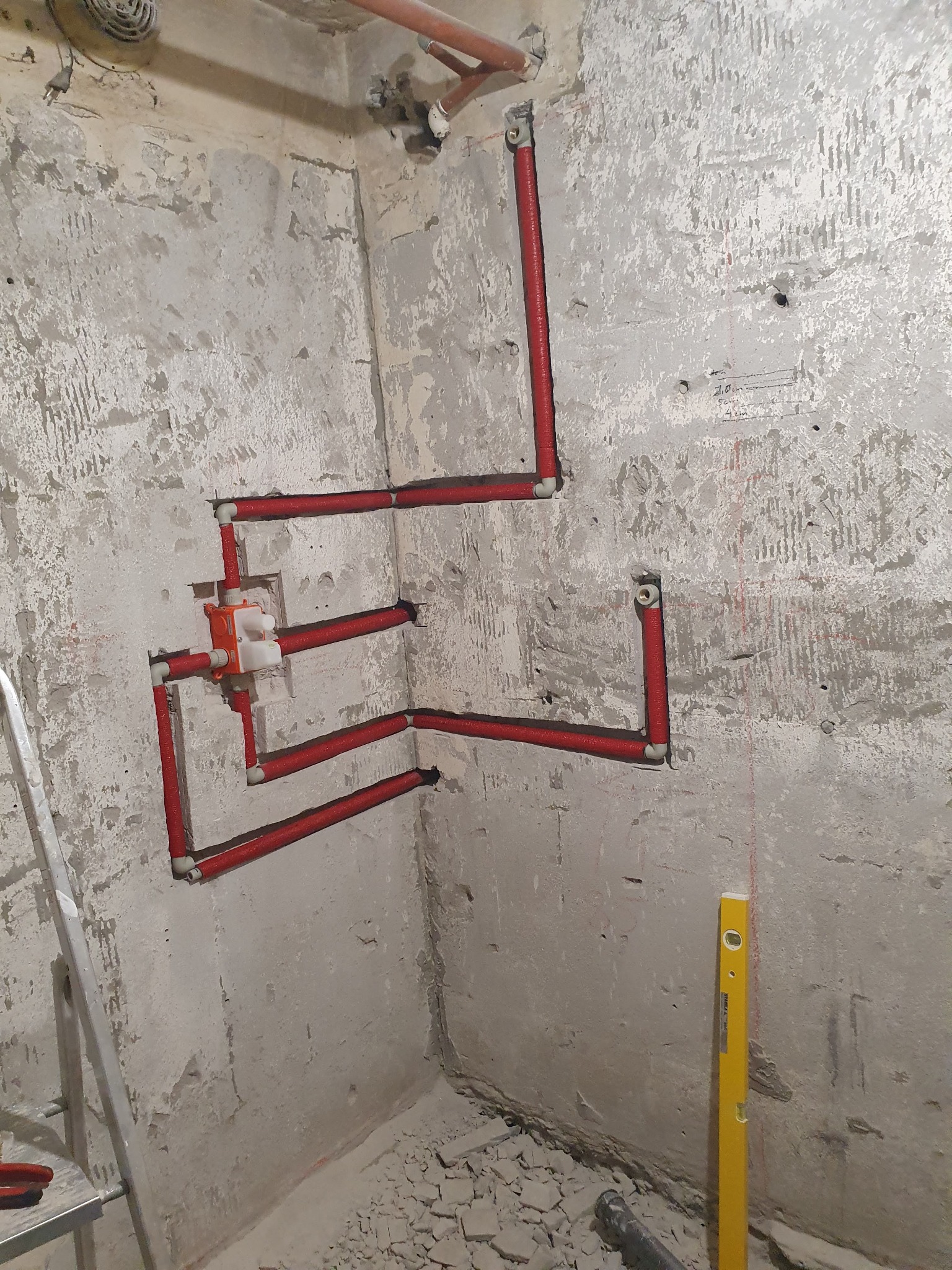 Surowe ściany w trakcie prac instalacyjnych, widoczne czerwone rury grzewcze ułożone na ścianie, żółta poziomica oparta o ścianę, fragment drabiny i gruz na podłodze.
