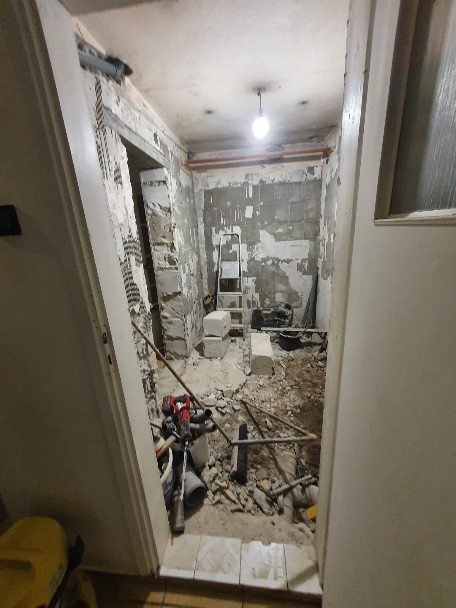 Wnętrze mieszkania w trakcie demontażu, widoczne fragmenty starej glazury na ścianach, gruz na podłodze, drabina oraz narzędzia budowlane, w tym młot udarowy i wiadro.