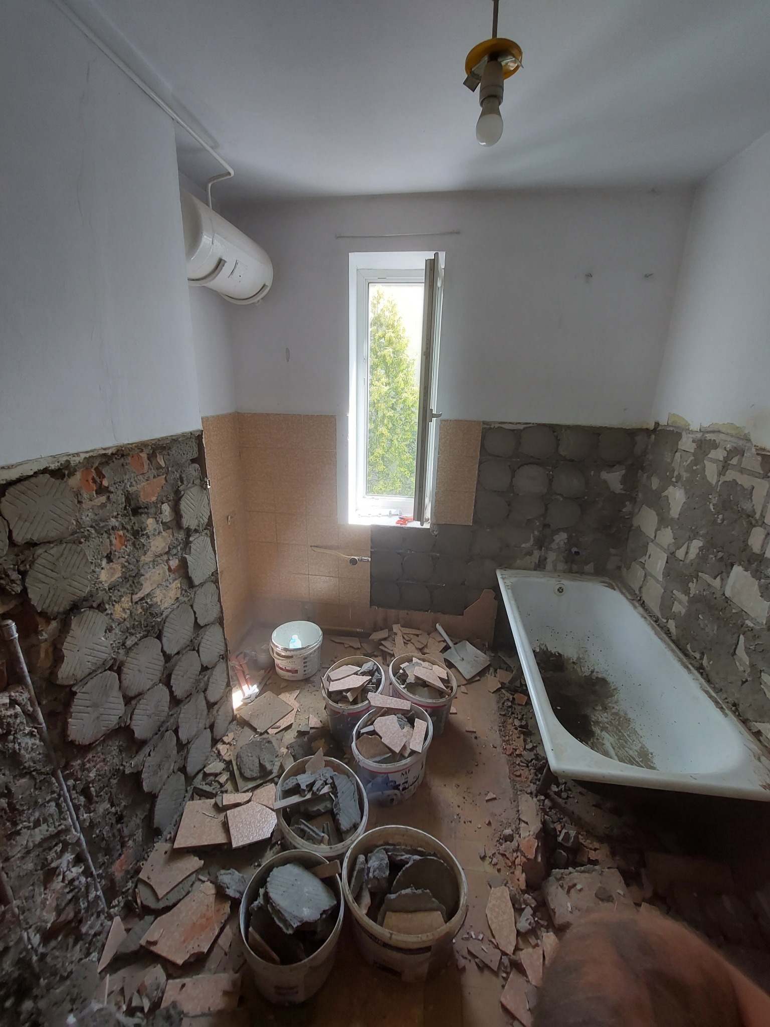 Demontaż płytek w łazience: widoczne fragmenty ścian bez płytek, gruz w wiadrach, brudna wanna, okno z widokiem na drzewa, stara lampa na suficie.
