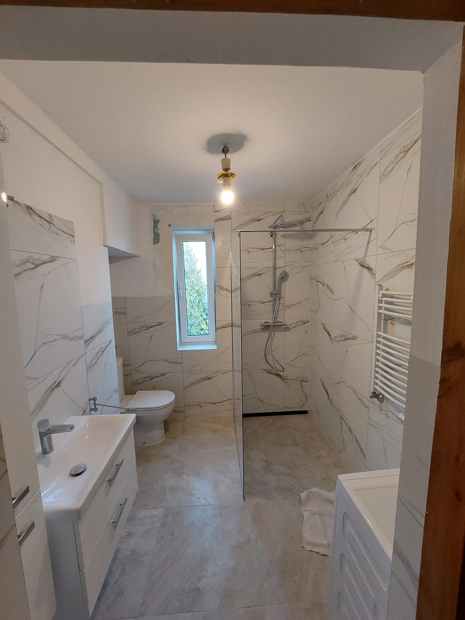 Wykończona łazienka z marmurowymi płytkami, kabiną prysznicową typu walk-in, umywalką z szafką i grzejnikiem drabinkowym; widoczny fragment ściany z widocznymi śladami po remoncie obok okna.