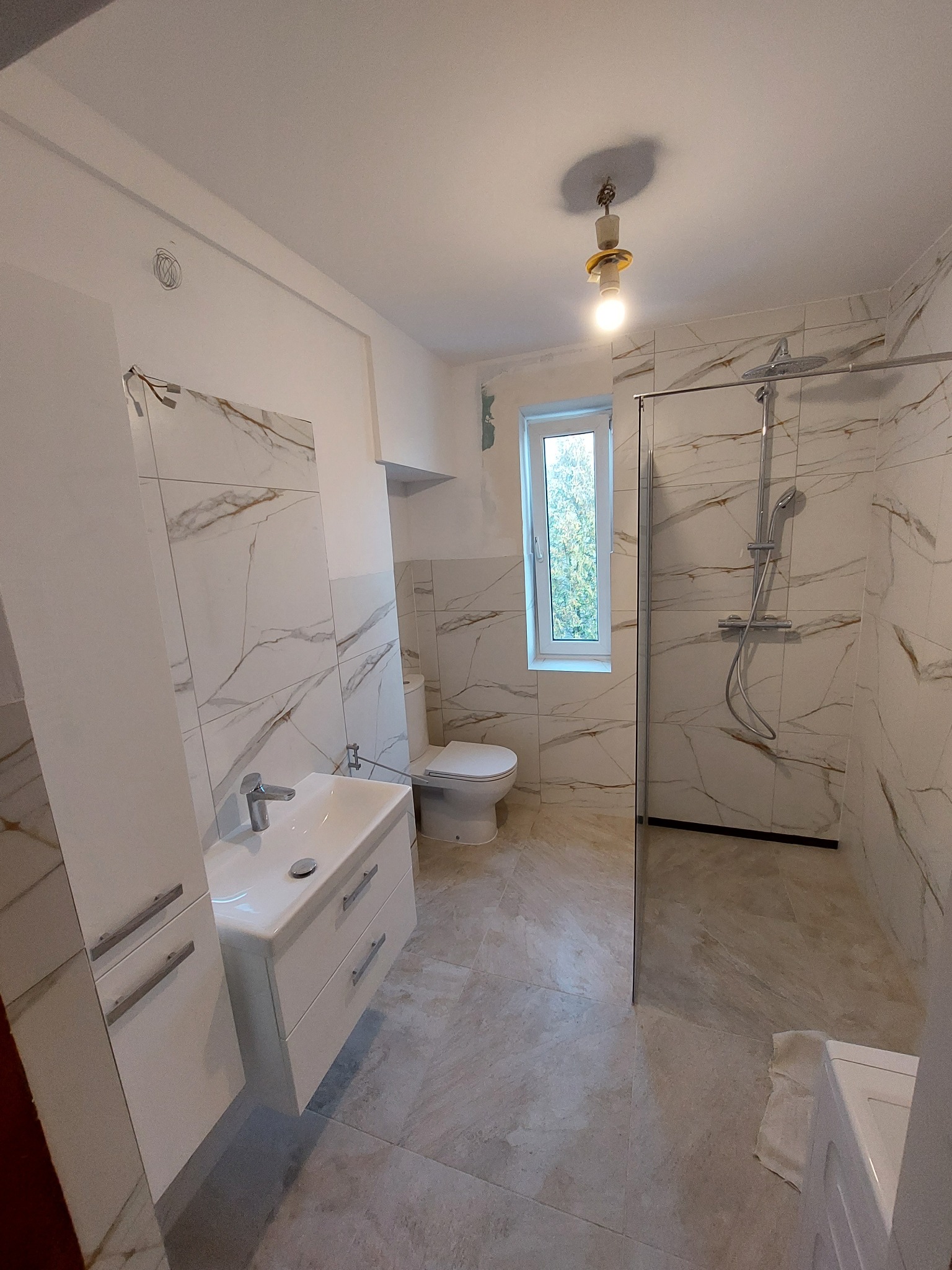Wykończona łazienka z kabiną prysznicową typu walk-in, umywalką z szafką i toaletą, wykończona płytkami imitującymi marmur z delikatnym złotym żyłowaniem, widoczna surowa żarówka na suficie.