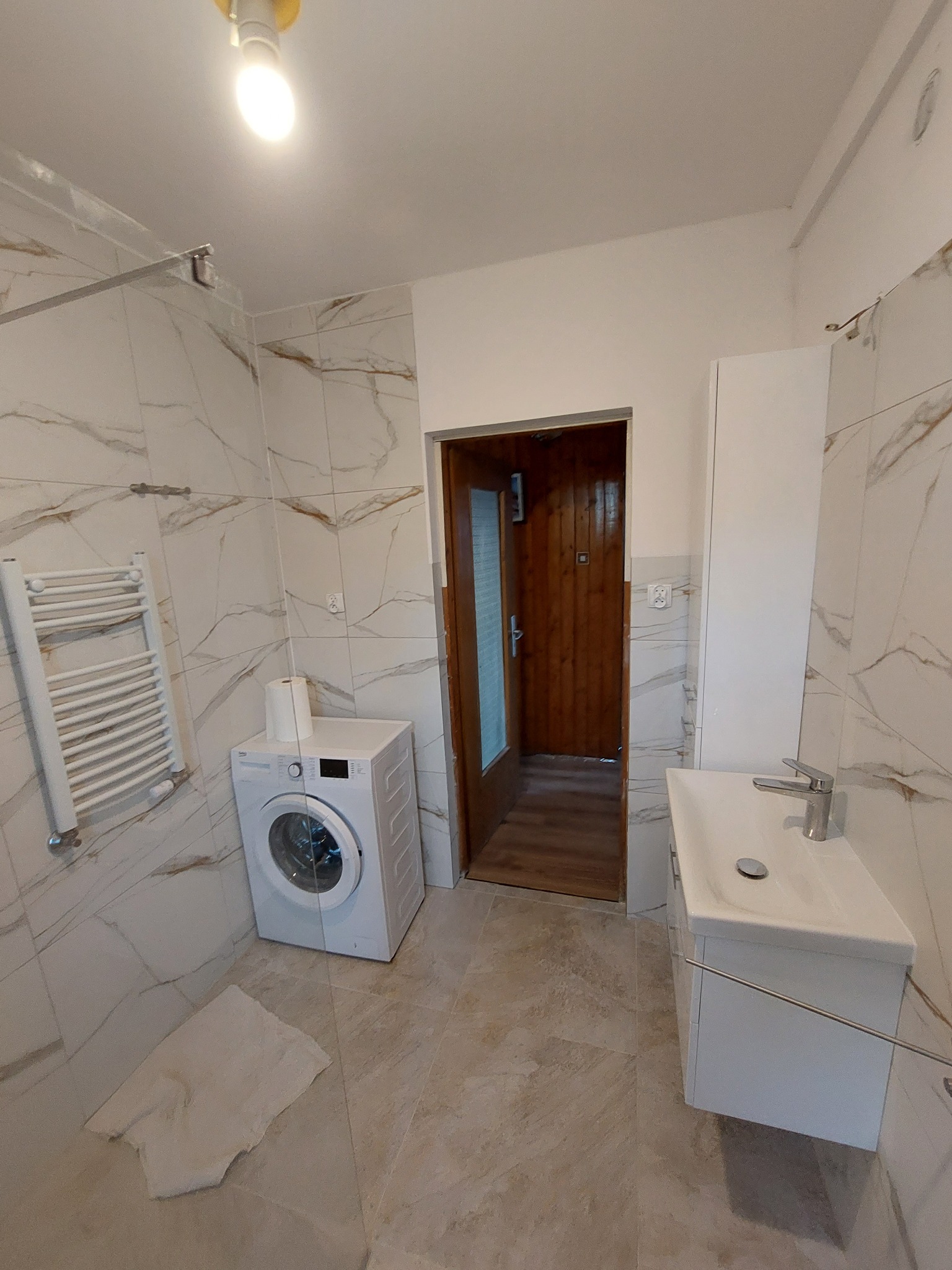 Wykończona łazienka z marmurowymi płytkami, białą pralką, grzejnikiem drabinkowym, szafką z umywalką i drewnianymi drzwiami z szybą.