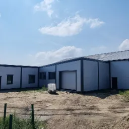 Nowoczesna hala magazynowa z szarej blachy trapezowej, widok z zewnątrz, jasne niebo z chmurami w tle, teren wokół niezagospodarowany.