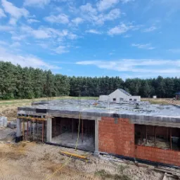 Budowa domu jednorodzinnego w stanie surowym otwartym, widoczne ściany z czerwonej cegły, betonowy strop z wystającymi prętami zbrojeniowymi, podpierające konstrukcje, na pierwszym planie ładowarka...