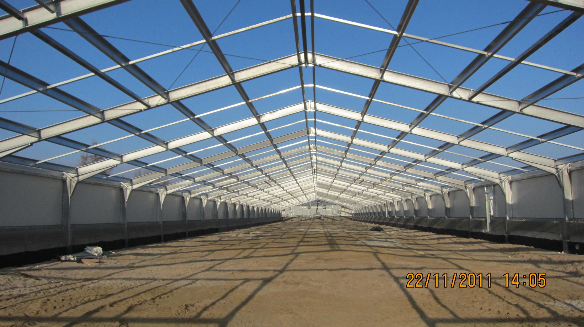 Wnętrze hali w trakcie budowy, widoczna stalowa konstrukcja dachu i ścian, ziemia na podłożu, jasne światło dzienne.