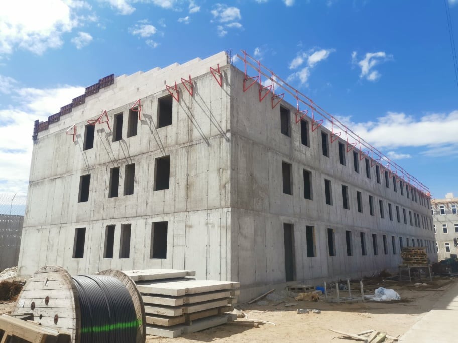 Surowy, trzypiętrowy budynek w trakcie budowy z widocznymi betonowymi panelami, niezabezpieczonymi otworami okiennymi i prowizorycznymi barierkami ochronnymi na dachu, na pierwszym planie zwój...
